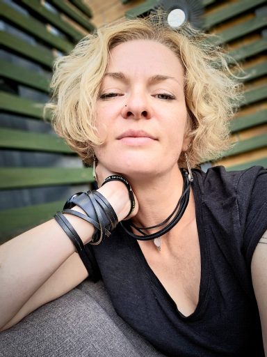 Gabi - Maker Behind Gabus81 Woman with short, curly blonde hair relaxing against a backdrop of green stripes.